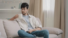 Millennial arabic guy indian handsome 30s man sitting at home on couch drinking hot tea coffee cappuccino cocoa from cup receive message looking at phone smiling chatting with friends with smartphone