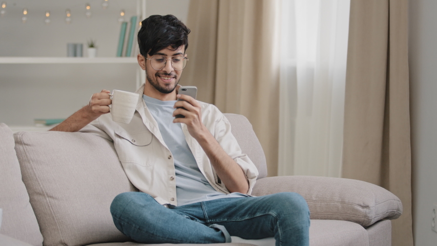 Millennial arabic guy indian handsome 30s man sitting at home on couch drinking hot tea coffee cappuccino cocoa from cup receive message looking at phone smiling chatting with friends with smartphone | Shutterstock HD Video #1087918179