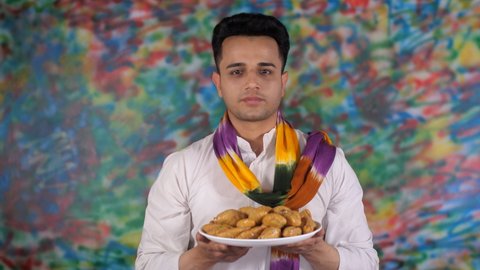 A cheerful Indian youngster greeting people on Holi with a plate of traditional Gujia. An attractive male wearing a white Kurta and a colorful Dupatta posing for the camera - festival of colors.
