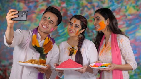 College friends wearing a white Kurta and a scarf enjoy clicking a selfie on Holi - festive season, togetherness and bonding. Cheerful siblings standing with a plate of Gulal and traditional Gujia.