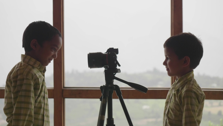 Two young cute Indian male school kids are playing with or making funny faces in front of a digital photography camera or DSLR with a background of misty mountainous valley out of window. | Shutterstock HD Video #1087940231