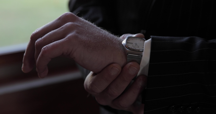 Groom in a black suit adjusts watch on wrist to straighten it out as he prepares for a wedding ceremony.
