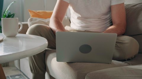 Young man using laptop typing and working sitting on the couch at home