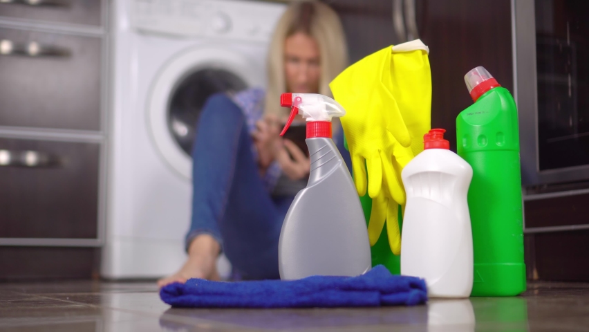 a happy modern woman using a home cleaning app on a smartphone in a modern home. She communicates in a messenger sitting by washing machine on floor. Bottles with the product are in foreground.