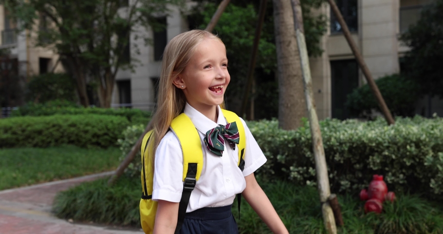 Happy little caucasian blonde girl seven years old in uniform with yellow backpack going back to school after summer vacations or Coronavirus covid-19 quarantine. 4k slow motion