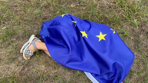 4k Little smiling girl covered with Eu flag lying on the lawn in the park.