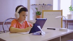A boy in white headphones is studying in an online school on a tablet