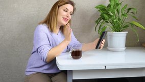 A happy young woman laughs, looks at the screen of a mobile phone, makes video calls on a mobile phone. The girl is sitting at the kitchen table and drinking coffee.