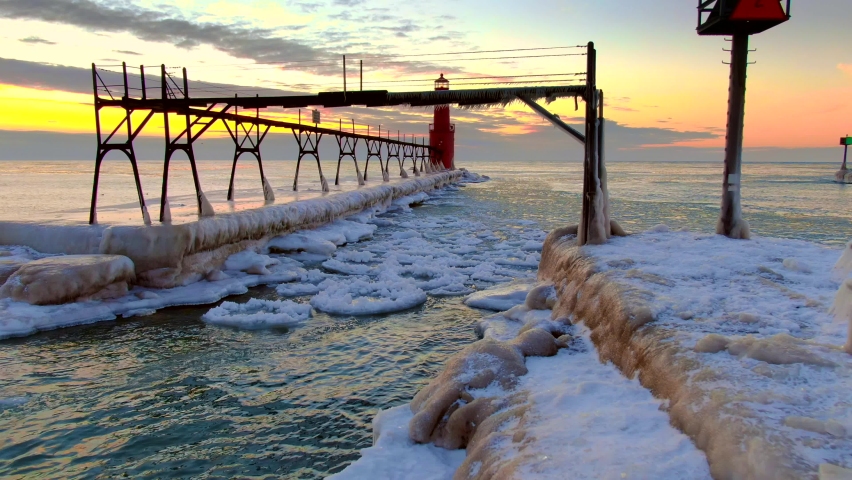 There is amazing beauty in this aerial view of an icy harbor in Winter, clouds on the horizon, waves reflecting the fiery sky, lighthouse beacon blinking, Lake Michigan in the Winter just before dawn.