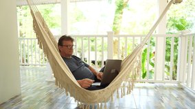 Senior man talking on internet via laptop in a hammock on a terrace home near tropical garden in island Koh Phangan, Thailand, close up