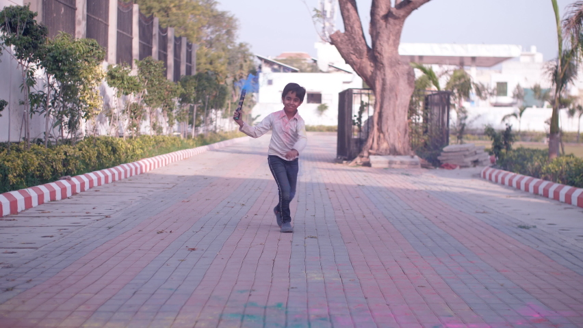 A young school-aged kid running on the pavement with a colored smoke stick - Holi celebrations, blue-colored smoke, creative, pyrotechnics. A smart little kid with Holi colors on his clothes and face.