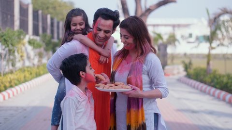 A young mother feeding Gujia to her family members - Holi celebrations, an Indian sweet, tasty cuisine. A small nuclear family with their faces smeared in Holi colors together in a public park.