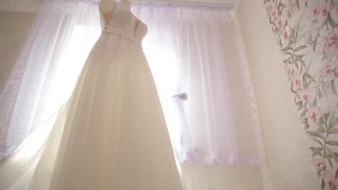 The wedding dress hangs on a mannequin, and is illuminated by the sun's rays from the window of the room.