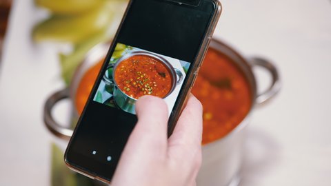 A Woman Looks Photo of Traditional Ukrainian Red Borscht in a Smartphone. Photographing food. Picture of food through the camera of a mobile phone in an application. Hands. Home kitchen. Lunchtime.