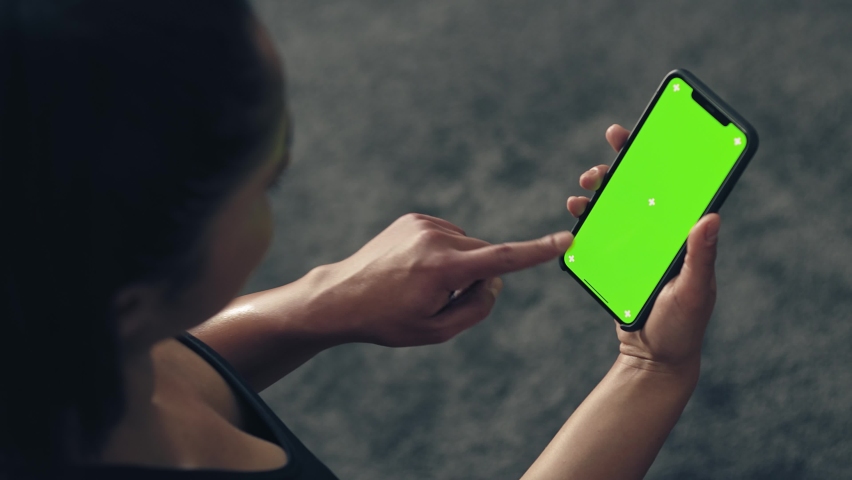 Back view of fitness woman using green screen smart phone at home. Woman preparing for workout using smart phone.Woman doing a tap on mobile phone while resting during fitness training