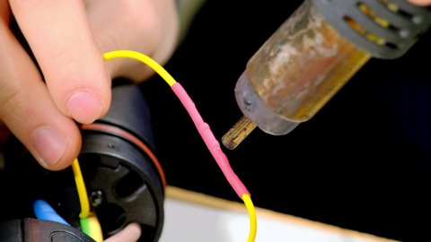 Foreman heats tube on wire to shrink using heat-gun at workshop. Specialist of maintenance prepapes plug cable for charging ev at table closeup