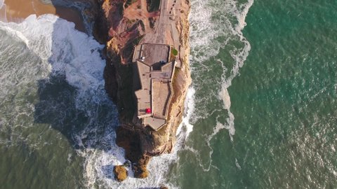 Dynamic Drone Aerial view of lighthouse on a cliff with a fortress on the coast of the Atlantic ocean in Nazare, or Nazare  Portugal, 4k , Red lighthouse  and gulf coastline in background.
