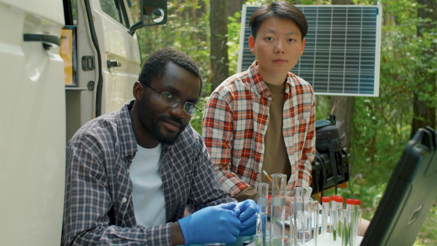 Medium portrait shot footage of two ethnically diverse scientists sitting against minibus in local forest doing chemical research