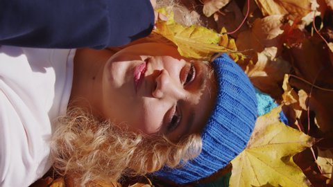 Vertical Video, Blonde Girl in Beanie Lying on Fallen Leaves, Cover Face With Leaf and Smiling, High Angle