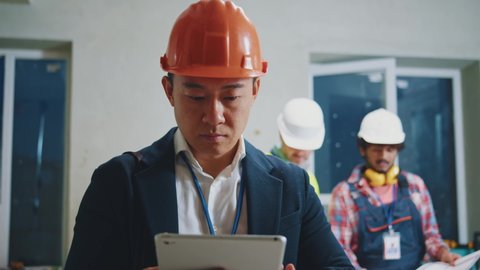 Portrait young asian engineer, architect wearing a white protective helmet in a suit use tablet computer stand. On bakground builders in hardhats working in building. Slow motion