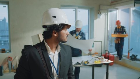 Portrait young engineer, architect wearing a white protective helmet in a suit look at camera smiling. On bakground builders in hardhats working in building. Slow motion