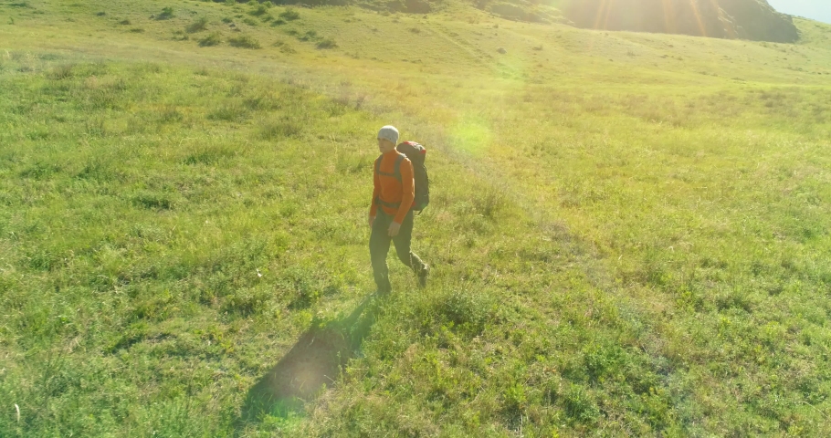 Aerial 4K UHD view. Low altitude flight over young man tourist walking across green mountain field. Huge rural valley and sunny meadow at summer day. Backpack hiking guy.