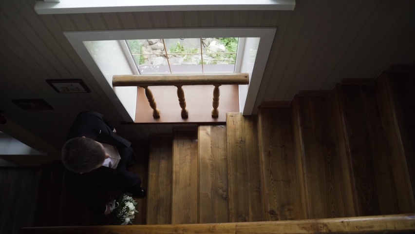 Groom in black suit and white shirt and black shoes with wedding bouquet in his hand climbs the wooden stairs to the houses