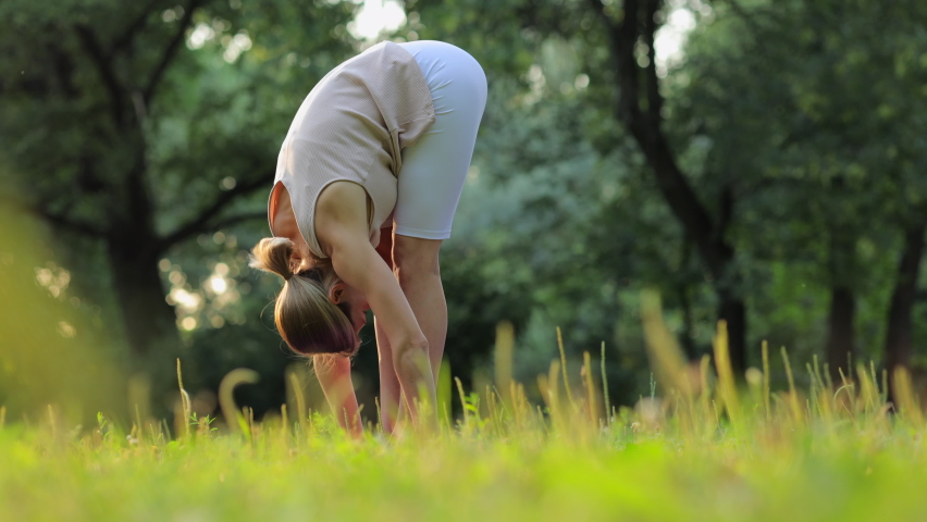 Young woman shaping body and exercising actively in green park on summer day spbi. 4k video Beautiful Caucasian female stretches arms, legs and looks with smile, she trains on grass on background of