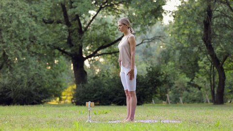 Young athletic female doing yoga online with device and standing in green park spbi. 4k video Beautiful caucasian woman stretches body and does asana in front of phone, joins hands in mudra and stands