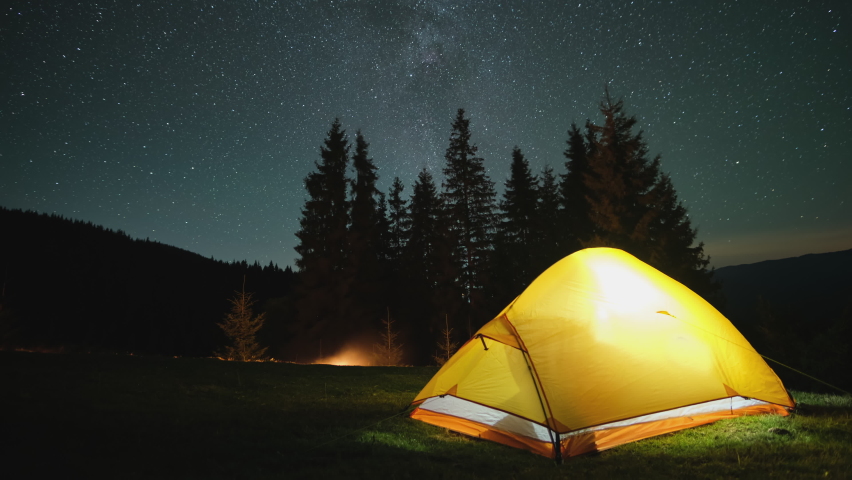 Time lapse of brightly illuminated camping tent glowing on campsite in dark mountains under night stars covered sky. Active lifestyle and traveling concept