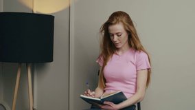 Young red-haired woman sitting on a chair isolated over white wall of the office, makes notes in a diary, smiling enjoying free time. Modern style. White wall.