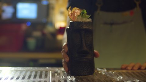 The bartender in the bar making the tiki style cocktail in the black tall special decorated glass. Shaking decorating and presenting the cocktail.