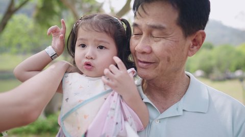 A happy healthy grandfather standing holding a little cute granddaughter inside an green fresh park, asian family spending outdoor leisure time together, a carefree lovely family happiness moments
