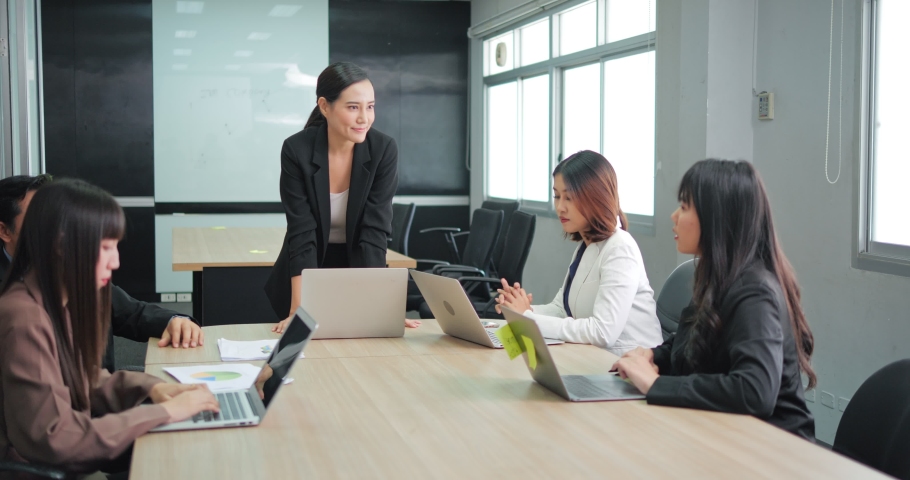 Confident Asian woman chairs a meeting with team in the office. Business woman stands to address meeting around board table. Royalty-Free Stock Footage #1087999047