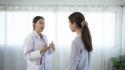 4K 50fps, white Asian female doctor standing and taking notes on a long-haired Asian patient wearing a long-sleeved shirt in a hospital examination room..