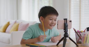 Asia cute smart boy alpha gen z kid enjoy fun hobby idea paint pencil color game on paper at online day care play school by vlog talk video call app. Young son study class tutor teach at home table.