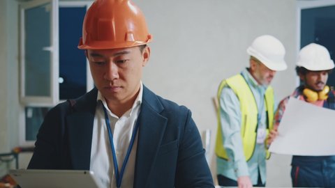 Close up portrait young asian engineer, architect wearing a white protective helmet in a suit use tablet computer stand look around. On bakground builders in hardhats working in building. Slow motion