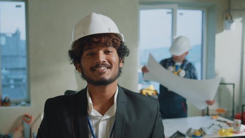 Happy portrait young engineer, architect wearing a white protective helmet in a suit look at camera smiling. On bakground builders in hardhats working in building. Slow motion