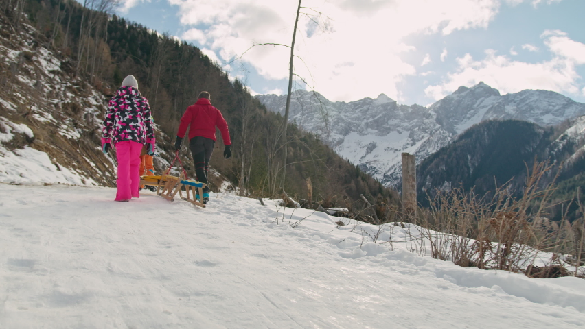 Family, a father and two children pulling sleds on a snowy track in a beautiful mountain landscape, tracking rear shot.