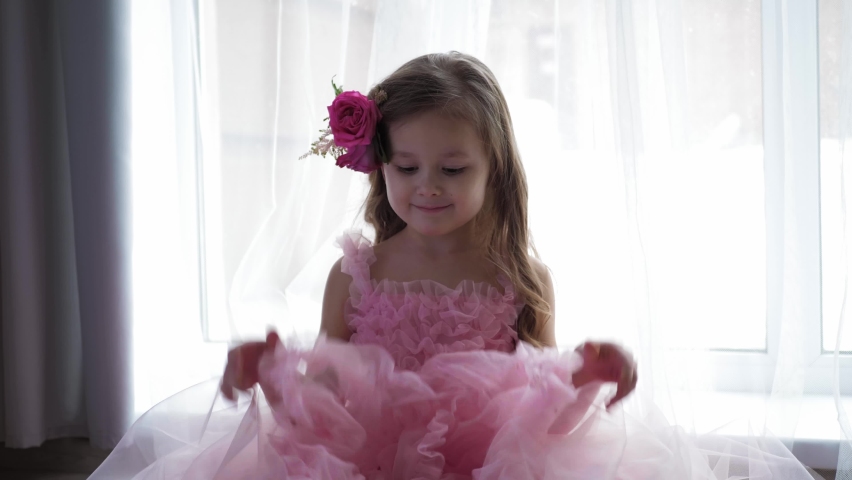 backstage of a photographer with a camera, beautiful portrait blonde of a girl child in a pink dress and with a rose flower in her head background of the window