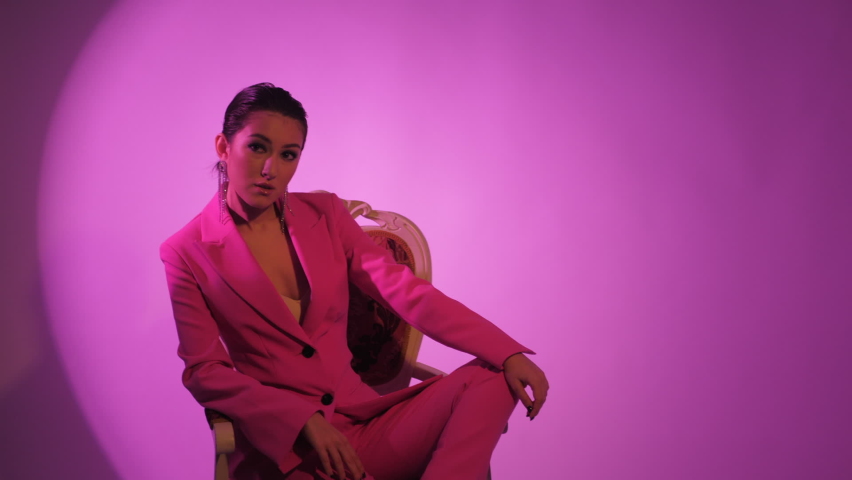 photographer backstage fashionable sexy woman model of Asian appearance in a business pink suit on the background in the studio and sitting on a chair
