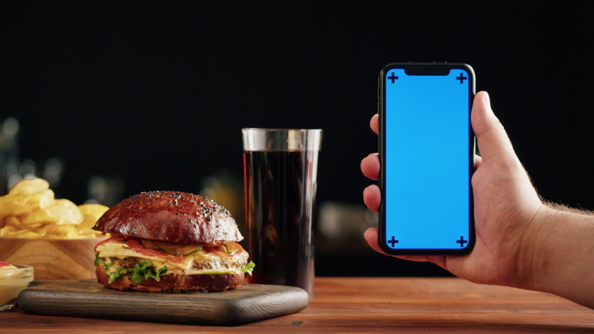 Tasty burger and smartphone with blue chroma key screen close-up, cooked juicy sandwich, fast food delivery application, recipe of preparing homemade hamburger with meat and fresh vegetables.