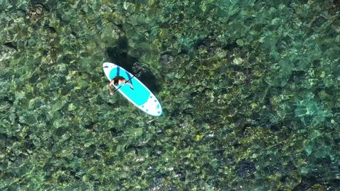 Top down girl rides a paddleboard on the crystal clear sea. Aerial view to woman on SUP board. Aerial view of young girl stand up paddling on vacation. Tracking shot of a young woman SUP boarding