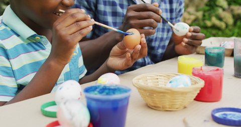 Animation of happy african american father and son painting easter eggs together in garden. family easter, celebration and spending quality time together outdoors.