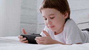 Little 5 year baby boy lays on bed and plays with smartphone. Closeup portrait. 4K video footage. Online education concept. White room and clothes. Child is holding and scrolling the telephone screen.