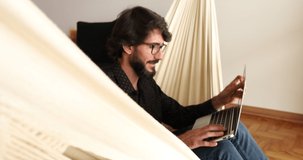 Young business man with black glasses working at home with laptop on a white hammock. Gray notebook for working. Home office concept. High quality video