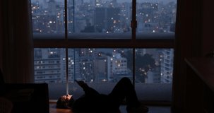 Young woman lying down on her apartment floor using a smartphone at night time with city view landscape in the background.  Mobile phone, technology, concept. High quality video