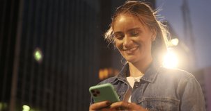 Young woman using a smartphone at night time with city view landscape in the background. High quality video. photo. Mobile phone, technology, urban concept