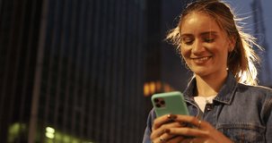 Young woman using a smartphone at night time with city view landscape in the background. High quality video. photo. Mobile phone, technology, urban concept