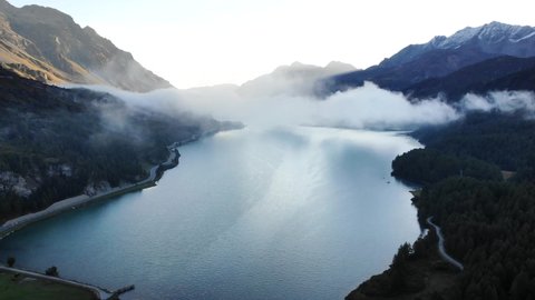 A sunrise aerial flyover through the clouds up above over Lake Sils in Maloja, Switzerland with a view of the peaks of Engadin and clouds moving over the water